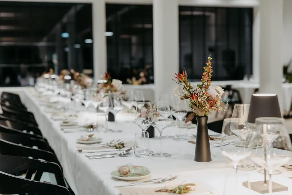Lange weiß gedeckte Tafel mit Blumen und Tischleuchten vor dunklen Fenstern