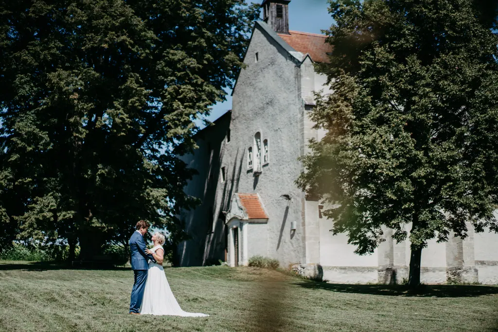 Brautpaar klein auf der Wiese vor einer alten Kirche zwischen hohen Bäumen