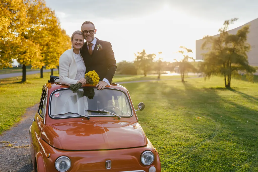 Brautpaar lehnt im Abendlicht an einem roten Oldtimer in der Herbstallee
