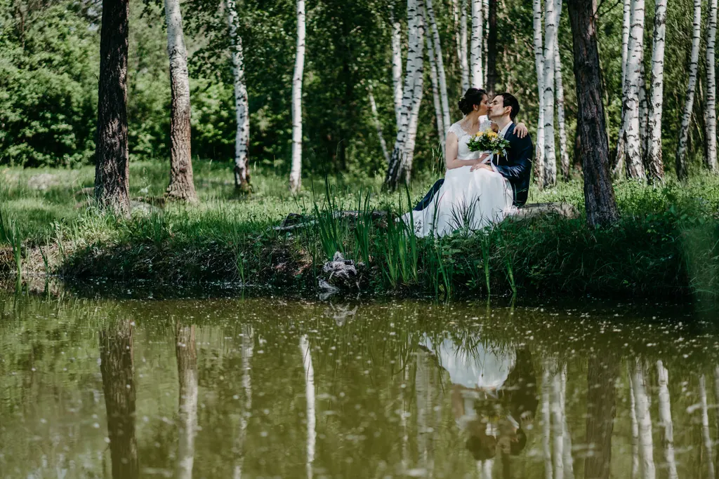 Brautpaar sitzt am Ufer eines Teichs zwischen Birken und spiegelt sich im Wasser
