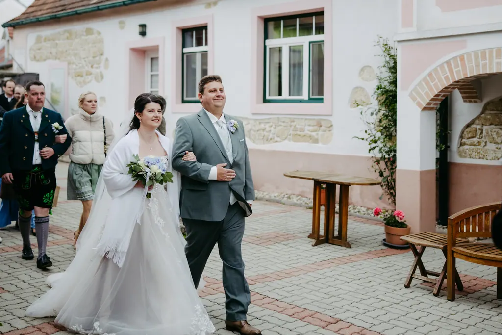 Brautpaar zieht untergehakt durch die Gasse, Gäste in Tracht dahinter