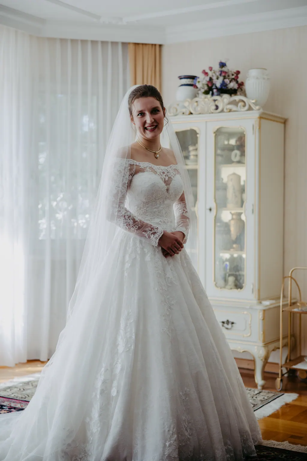 Braut im Spitzenkleid mit Schleier steht lächelnd im hellen Wohnzimmer