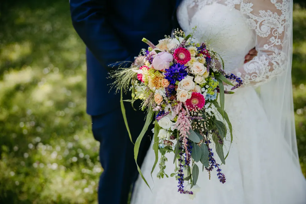 Bunter Brautstrauß mit Rosen, Gerbera und Gräsern vor dem Spitzenkleid