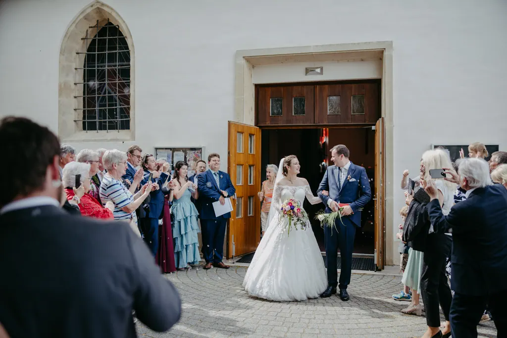 Brautpaar tritt aus dem Kirchenportal, Gäste stehen links und rechts Spalier