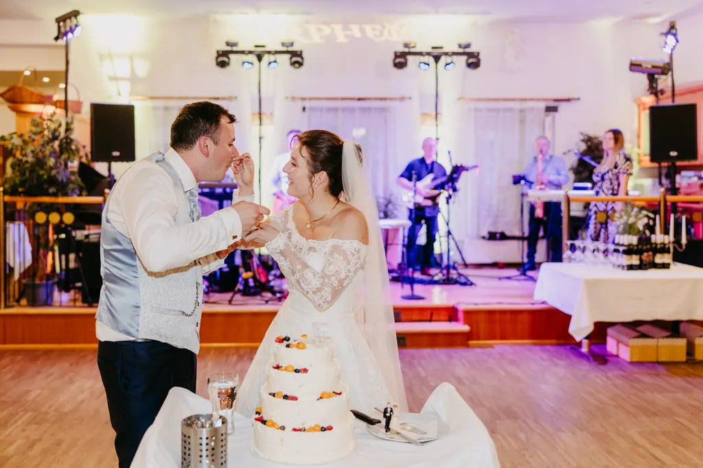 Brautpaar füttert sich gegenseitig mit Torte, im Hintergrund die Band