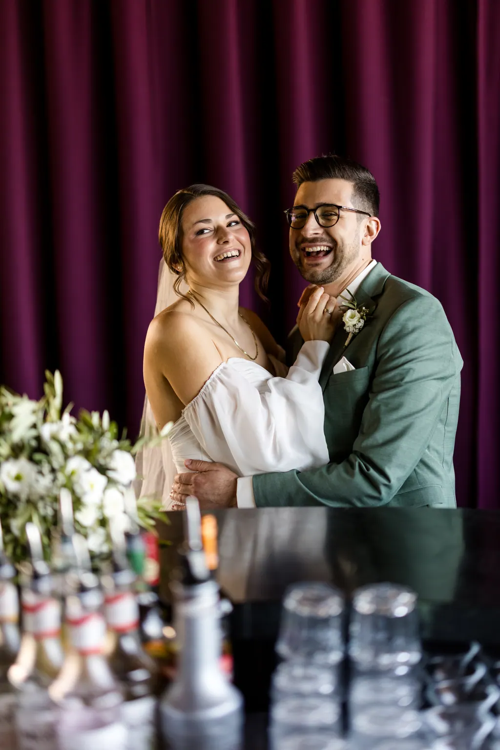 Brautpaar lacht ausgelassen an einer Bar vor violettem Vorhang