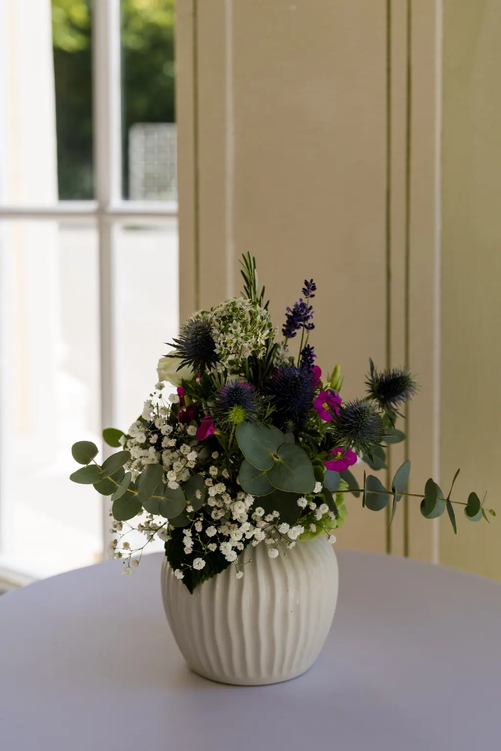Vase mit Wiesenblumen und Disteln auf weißem Tisch vor hohem Fenster