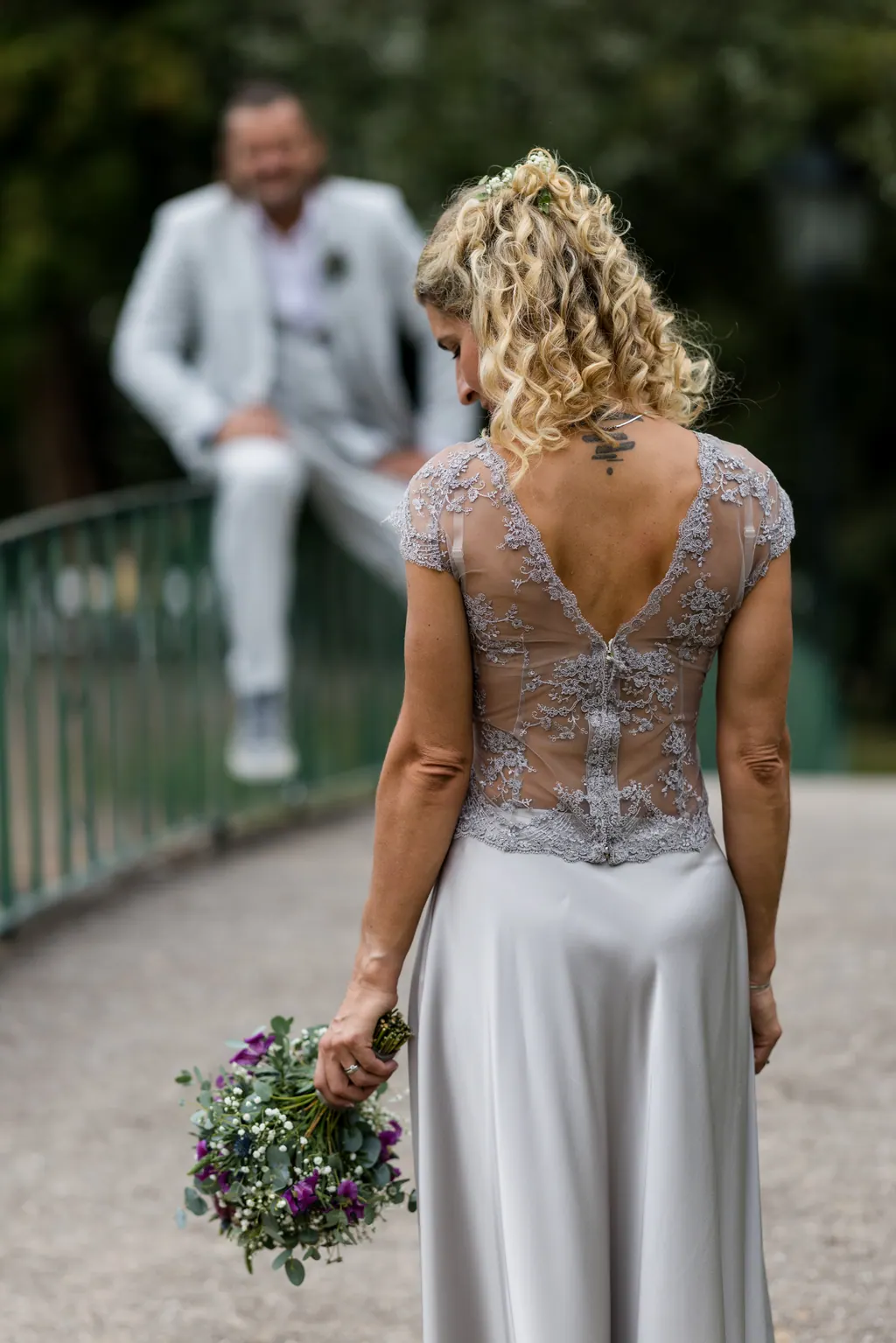 Braut im Spitzenrücken-Kleid von hinten, Blick zum Bräutigam auf der Brücke